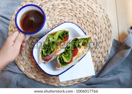 a plate of scrambled egg and vegetable pita sandwich on the breakfast table with a hand holding a mug of black tea