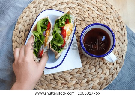 a hand holding a scrambled egg and vegetable pita sandwich on the breakfast table with a mug of black tea