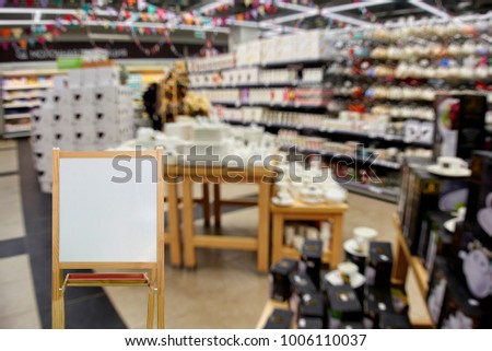 Chalkboard in wooden frame as place for your text on the foreground.  The department of dishes, shelves and racks with plates, mugs, vases, out of focus, blurred, in the background.