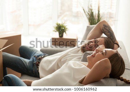 Happy couple relaxing on sofa having fun on moving day, excited young homeowners enjoying relocation in into new home, positive laughing man and woman sitting on couch with packed cardboard boxes Royalty-Free Stock Photo #1006160962