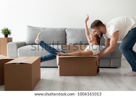 Happy couple having fun laughing moving into new home, young excited woman riding sitting in cardboard box while man pushing it, cheerful roommates playing while packing unpacking belongings together Royalty-Free Stock Photo #1006161013