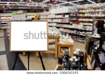 Chalkboard in wooden frame as place for your text on the foreground. The department of dishes, shelves and racks with plates, mugs, vases, out of focus, blurred, in the background.