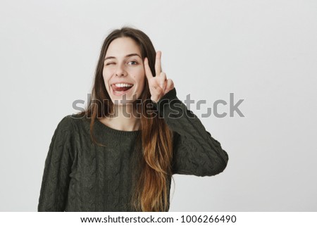 Portrait of cute attractive young caucasian girl winking and sticking out tongue while smiling broadly and showing victory gesture, isolated over white background. She likes japanese cartoons.