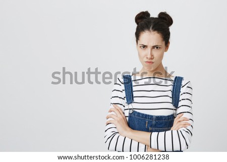 Picture of funny brunette girl in denim overall posing at studio, keeping arms folded, having offended look, pouting lips, showing her dislike, negative attitude and reaction.