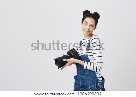 Positive cheerful carefree female photographer with two hairbuns wearing denim overall using modern camera. Pretty girl with smile holding photo camera, posing with joy against gray background