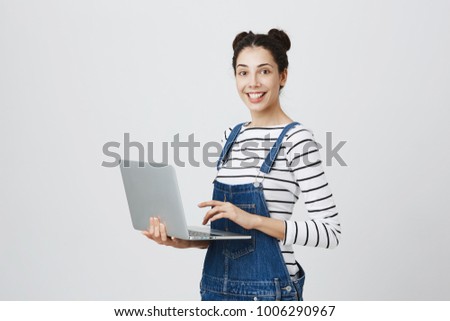 Positive young caucasian brunette female copywriter with two hairbuns in denim clothes using modern generic laptop computer and smiling cheerfully. Modern technologies. Advertising concept