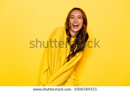Excited young woman with curly hair in yellow sweater, widely smiling, looking at camera. Isolated on yellow background.