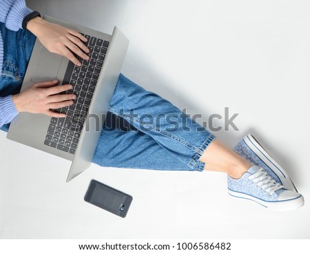 The concept of blogging, working on a laptop. Using a laptop sitting on a white floor. Modern technologies. Female legs in jeans and sneakers. Top view. Flat lay. Royalty-Free Stock Photo #1006586482