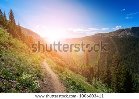 Dirt path hiking trail climbs through the Colorado mountains with the colorful light of the bright sun shining over the distant horizon