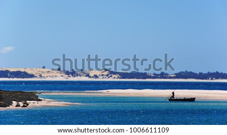 white beaches and blue water of the Bazaruto Islands, on the water a boat with two fishermans, in the background hilly and sandy coast of Mozambique, all this under a blue and empty sky               