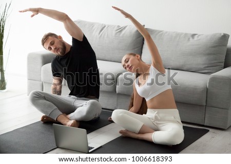 Sporty young couple warming up stretching watching fitness video tutorial online on laptop, fit man and woman doing workout at home sitting on mat in living room practicing yoga side bending exercise Royalty-Free Stock Photo #1006618393