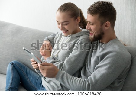 Smiling young couple embracing holding tablet looking at screen together sitting on couch at home, happy man and woman watching interesting video movie online or making internet call, reading news Royalty-Free Stock Photo #1006618420