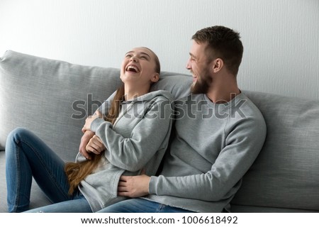 Young happy couple having fun talking laughing relaxing at home on couch, boyfriend embracing girlfriend telling funny joke sitting on sofa, humor in relationships, enjoying weekend together