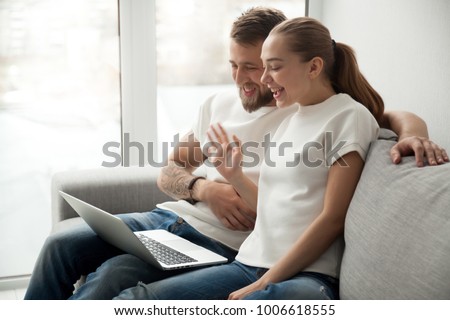 Smiling young couple making distance video call on laptop sitting on sofa at home, happy family calling friends online, cheerful man and woman waving hand looking at screen at web camera greeting Royalty-Free Stock Photo #1006618555