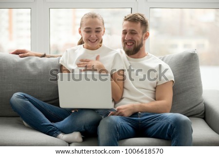 Young happy couple laughing with joy sitting at home on sofa looking at laptop screen, cheerful man and woman watching funny video on computer, having fun enjoying positive amusing content online
