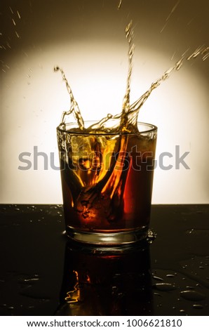 Glass of whiskey with spray from the thrown ice / In the picture there is a glass of whiskey with ice, standing on a table filled with liquid