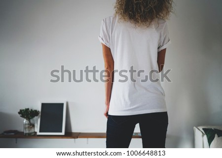 rear view of a sporty young hipster girl with light curly hair dressed in a blank white T-shirt and blue skinny jeans. Empty space for logo and design. Horizontal mockup. White wall background.