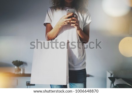 Young hipster girl is holding a blank white paper package in her hands with an empty space for logo and design.Cropped photo of a girl who writes a text message on a modern mobile phone. Bokeh light