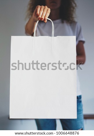 Young hipster girl wearing blank white t-shirt and holding white paper package with empty space for your logo or design, mock-up of shopping bag with handles. Vertical photo.