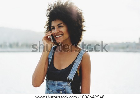 Attractive smiley mixed race girl talking on her mobile phone with her friend in the rays of the evening sun near the sea. Young woman wearing in a casual clothes using cellphone for business call.