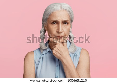 Headshot of grey haired senior female thinks about retirement, looks confidently, keeps hand under chin, feels lonely and bored on pension, needs communication and support. People and age concept