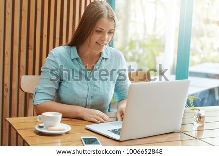 Photo of young pleasant looking female copywriter being busy with searching inforamtion on different websites, spends time at coffee shop, enjoys hot tasty latte, makes plans for coming weekends