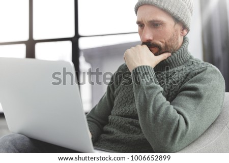 Serious concentrated unshaven male looks attentively into laptop computer, books on internet websites or watches training webinar, sits in coworking space. Stylish businnessman keyboards information