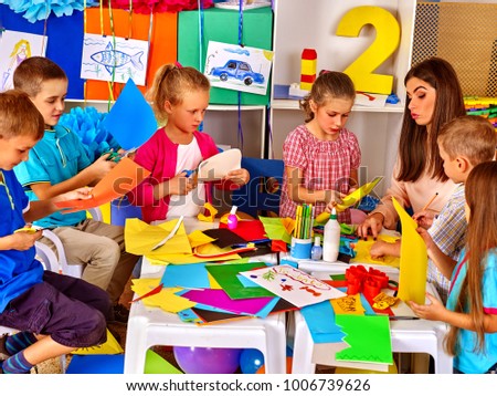 Children and colored paper. Making something on table in primary school. Craft lesson in primary school. Development kids craft at class in school. Children with teacher make overall project.