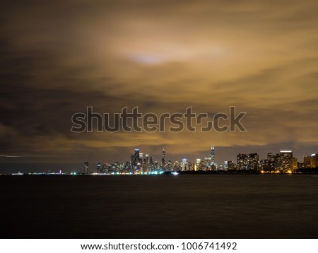 Beautiful night time long exposure photograph of the colorful lights of the buildings in the Chicago city skyline with Lake Michigan in the foreground and cloudy sky above just after sunset.