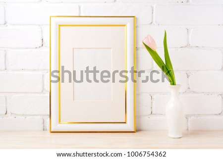 Gold decorated frame mockup with soft pink tulip in elegant vase near white painted brick wall. Empty frame mock up for presentation artwork. Template framing for modern art.