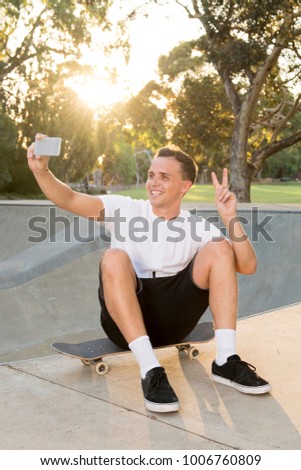 young happy and attractive American man 30s sitting on skate board after sport boarding training session taking selfie photo portrait or picture on mobile phone on sunset half pipe track park
