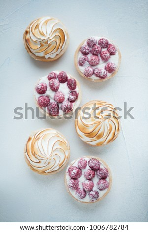 Delicious lemon and raspberry tartlets with meringue on a white vintage plate. Sweet treat on a light blue background. Flat lay and copy space. Top view