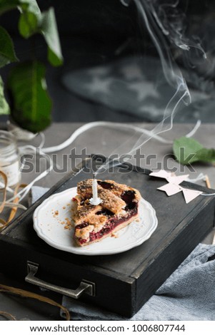 A piece of a birthday cherry cake with a burning one white candle. Atmospheric beautiful food birthday photo with home cherry pie, flowers, ribbons and decoration.