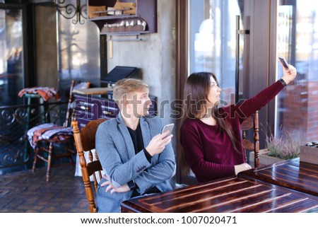 Physicist with smartwatch on hand in cafe with colleague taking selfies using smartphone wait friend, irritated guy checking social networks watching smiling Asian girl. Young people sitting near