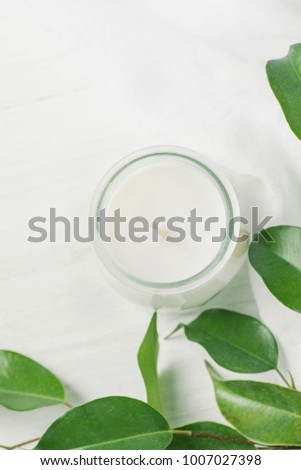 White Candle in Glass Jar Ficus Tree Branch with Young Fresh Green Leaves on White Wood Background. Styled Stock Photo for Social Media Blog. Wellness Spa Purity Tranquility Concept. Copy Space