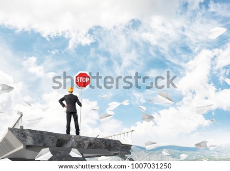 Rear view of engineer in helmet holding stop sign while standing among flying paper planes on broken bridge with skyscape on background. 3D rendering.