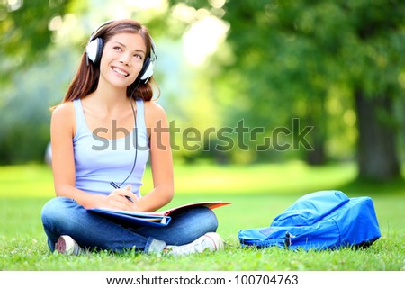 Female student girl outside in park listening to music on headphones while studying. Happy young university student of mixed Asian and Caucasian ethnicity.
