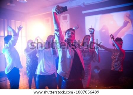 Crowd of trendy young people dancing in nightclub and enjoying party, focus on  handsome young man taking selfie on dance floor