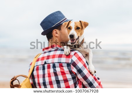 Small beautiful dog on owner`s hands. Stylish young male model dressed in fashionable clothes carries his pet and bag, stand at seashore, come to admire wonderful landscapes and breath fresh air Royalty-Free Stock Photo #1007067928