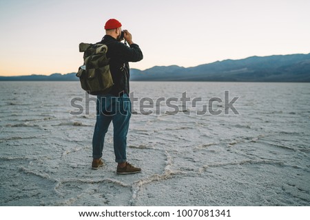 Hipster guy traveling in wild environment of lowest point if America using camera for making photos of landscape, back view of skilled photographer taking picture exploring arid lands of Badwater