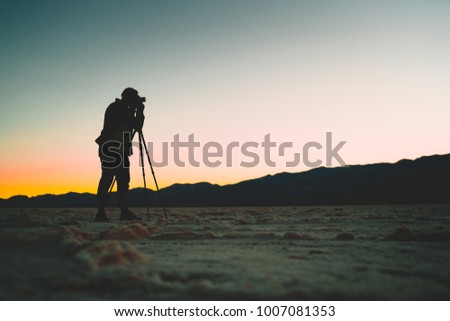 Silhouette of professional adventures photographer taking photo of beautiful sunset in Death Valley on camera with tripod equipment, man traveler shooting colourful skyline scenery view in the dusk