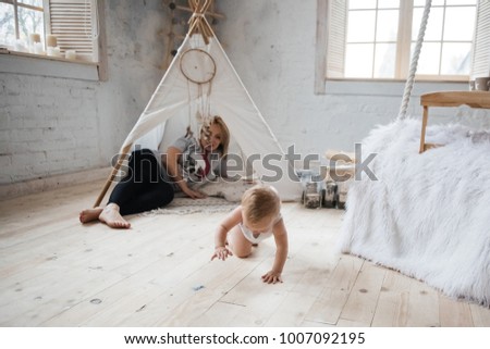 Mom and son with blond hair near the window. A room in Scandinavian style. They embrace, they play in their pajamas. Suspended bed. children's wigwam.