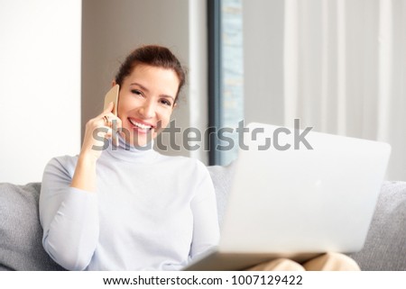 A happy woman using her cell phone and making call while sitting on sofa with laptop and working from home. 