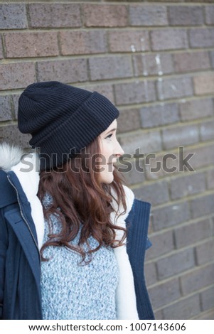 young woman in dark hat and coat leaning casually against a brick wall smiling. casual and fun. 