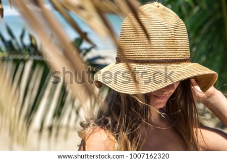 beautiful girl in a summer hat on a background of palm leaves on the beach, face covered with hat, closeup, holiday concept