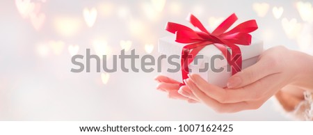 Valentine gift. Beauty Woman hands holding Gift box with red bow over holiday  background with glowing hearts bokeh, close-up. pastel colors. Wide angle format backdrop