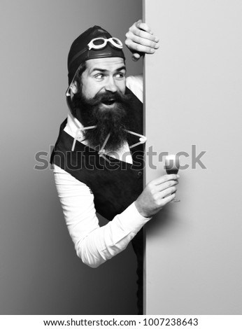 handsome bearded pilot man with long beard and mustache on funny face holding glass of alcoholic shot in vintage suede leather waistcoat with hat and glasses on blue green studio background