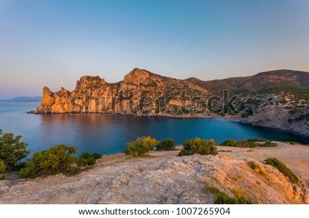 View of Black Sea bay with rocky shore lit with low sunrise light, stony path with bushes and grass in front and far mountains on horizon at sunrise in Crimea