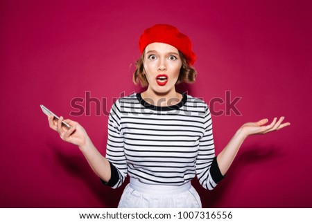 Confused ginger woman holding smartphone and looking at the camera over pink background