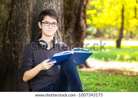 education portrait young, handsome and smart man asian student reading book, textbook, for exam and listen music by earphone from tablet with happy, smile, mood and relax in park with blur copy space 